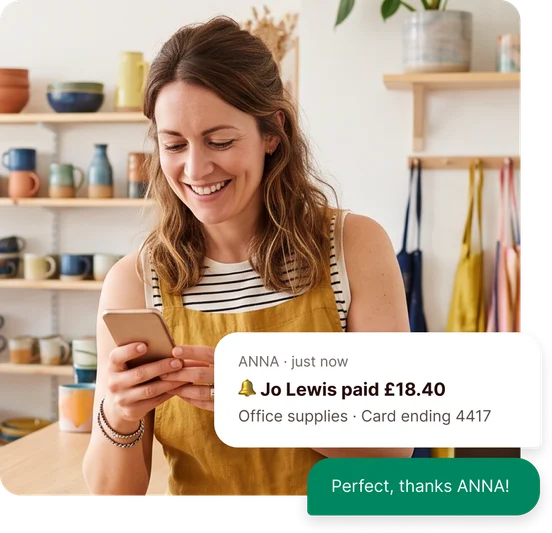 Smiling shop owner reading a phone in a pottery shop, with an ANNA notification that Jo Lewis paid £18.40 for office supplies on card ending 4417 and the reply “Perfect, thanks ANNA!”