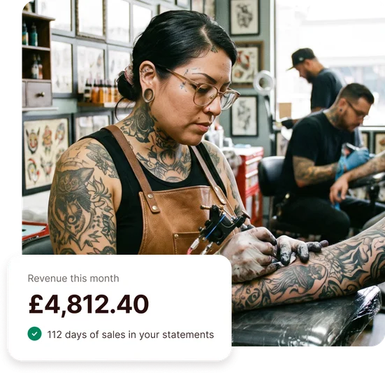 Tattoo artist working on a client’s arm in her studio, with a card showing £4,812.40 of revenue this month and 112 days of sales in her bank statements